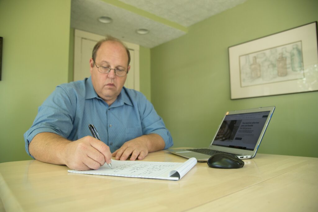 A man with glasses writes notes in a notepad at a wooden table, with a laptop displaying a webpage beside him, in a green-lit room.
