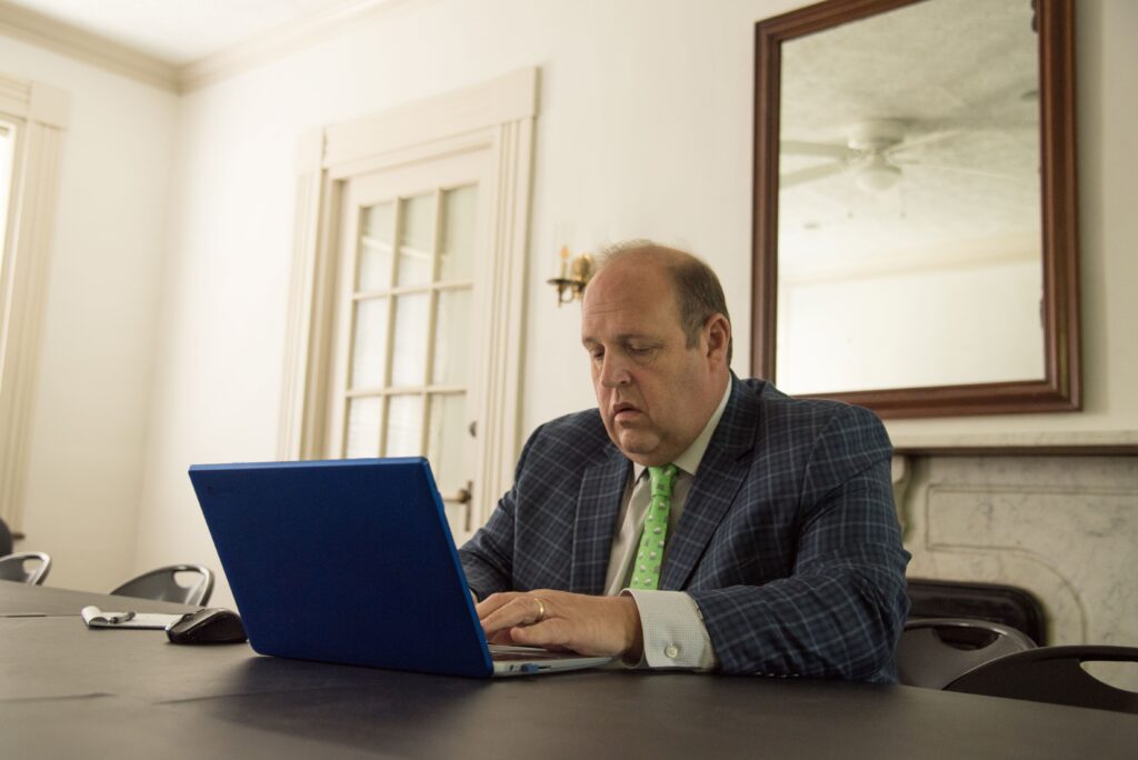 A focused man in a suit works on a blue laptop at a conference table, surrounded by a bright, minimalist meeting room.