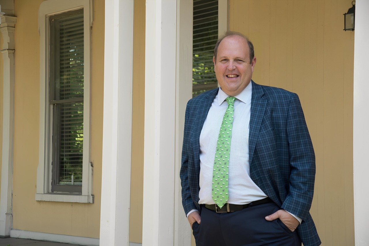 Smiling man in a checked blazer and green tie stands by a yellow wall with white pillars. The image conveys a welcoming and professional atmosphere.