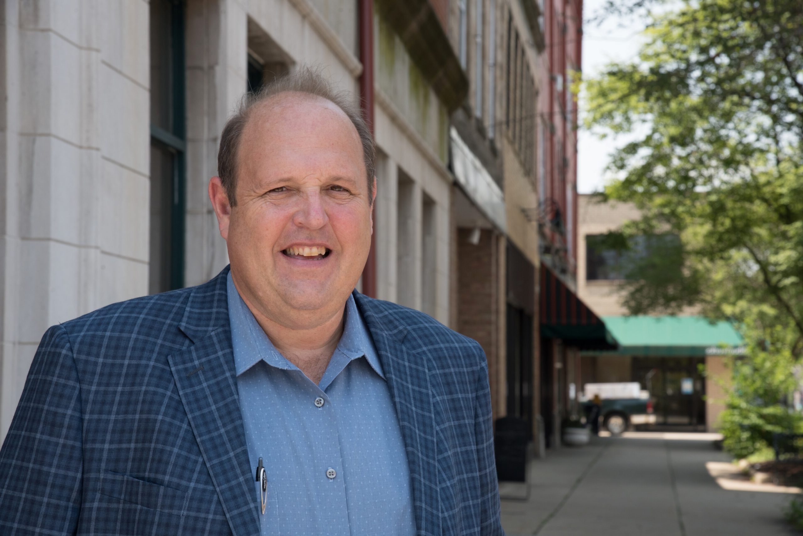 Smiling man in a blue plaid blazer stands on a sunny street, framed by buildings and greenery, suggesting a friendly community atmosphere.