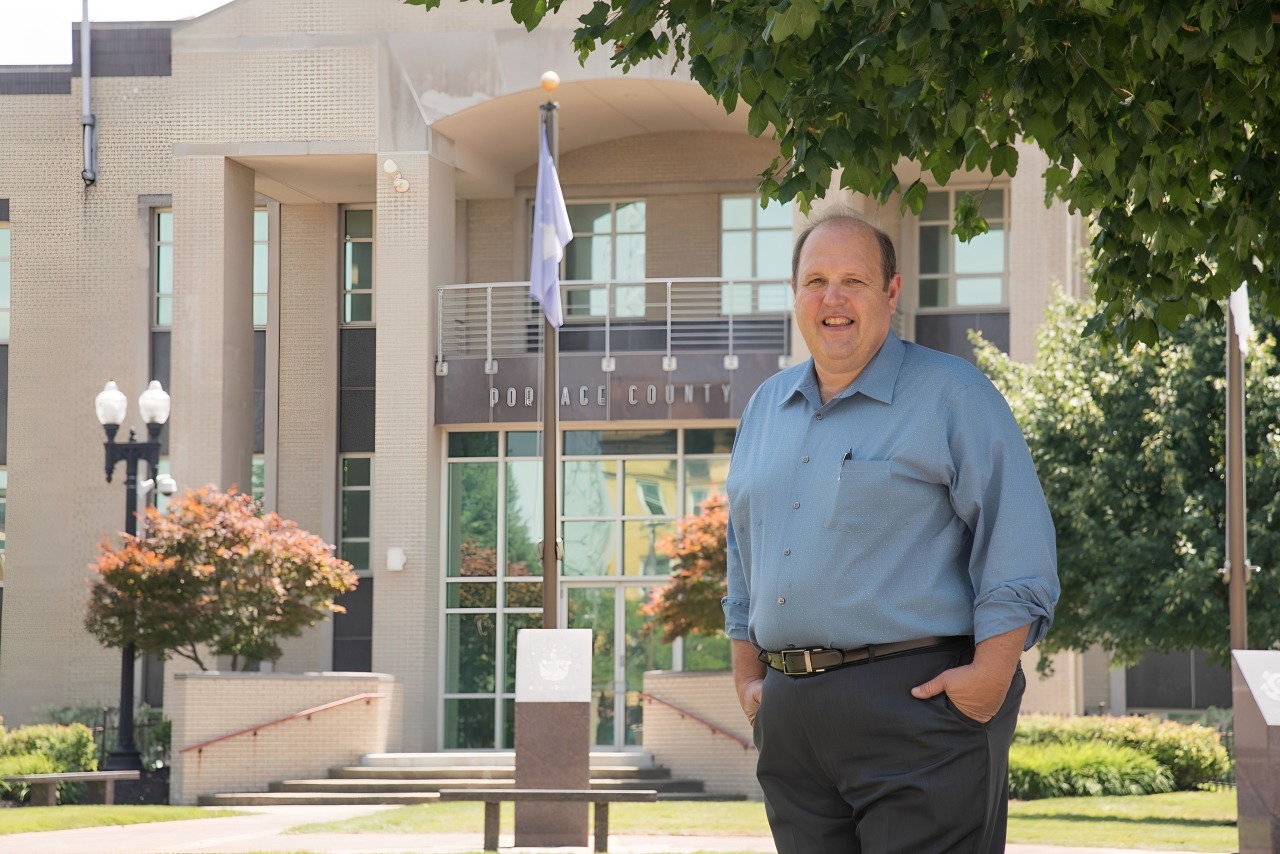 Smiling man in a blue shirt stands outside the Portage County government building, showcasing local leadership in a community setting.
