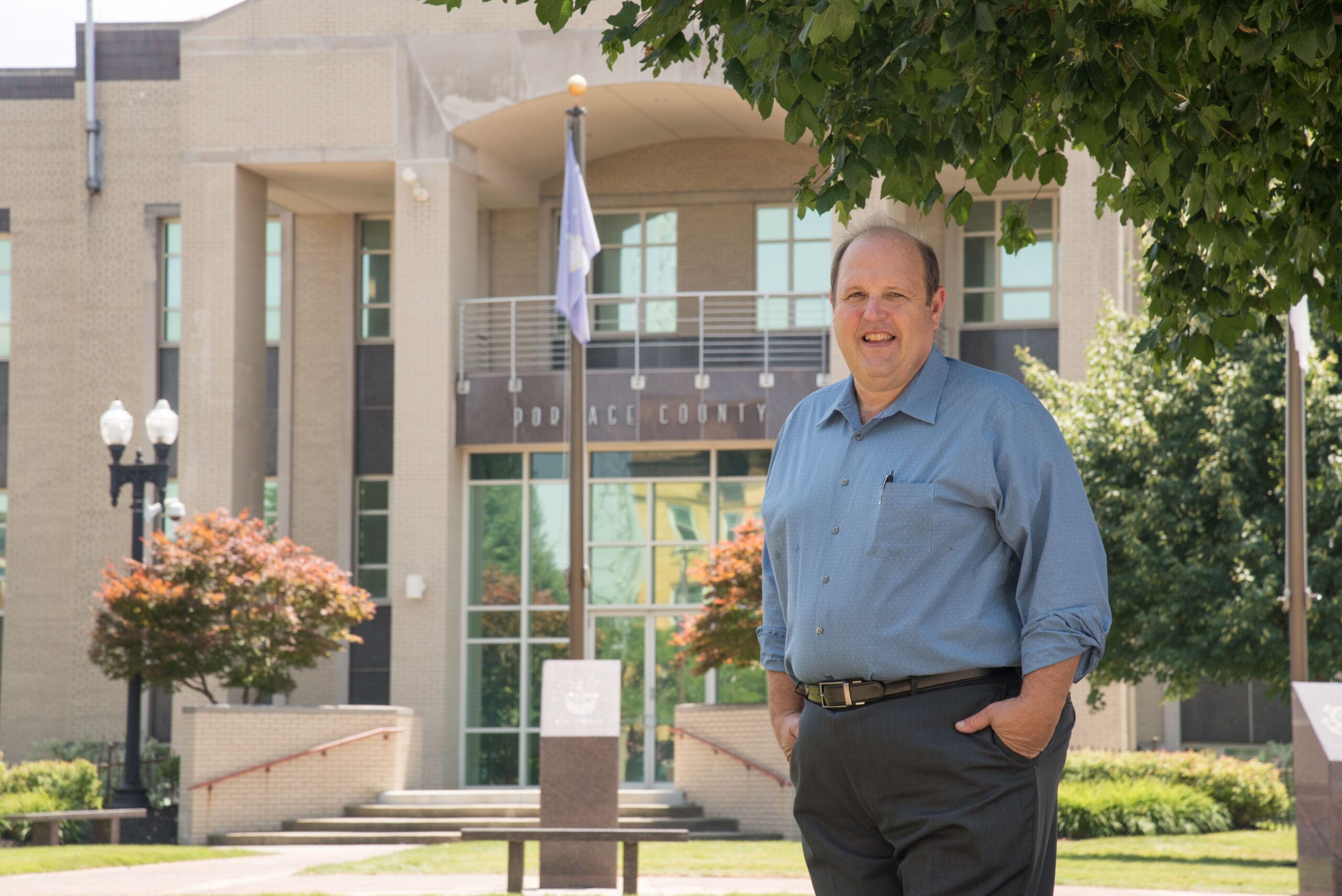 Smiling man in a blue shirt stands outside the Portage County government building, showcasing local leadership in a community setting.